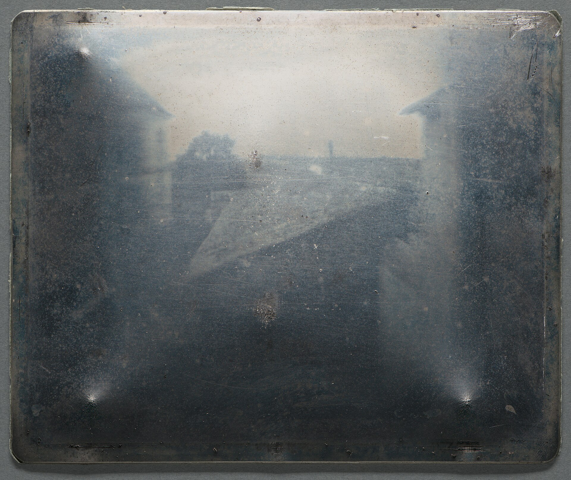 Fotografia d&rsquo;una placa metàlica amb una imatge a sobre d&rsquo;uns edificis. És &ldquo;Vista des de la finestra a Le Gras&rdquo;