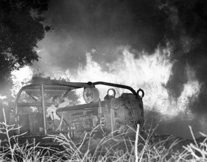 Fotografia en blanc i negre. Un home intenta fer un tallafocs amb una màquina mentre les flames son darrere seu